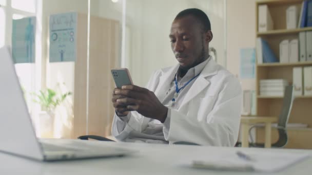 Beyaz Önlüklü Siyahi Erkek Doktorun Klinikteki Masasında Otururken Akıllı Telefondan — Stok video