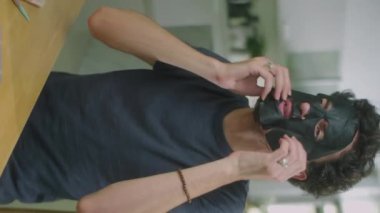 Erkek güzellik blogcusunun evde kozmetik ürünler incelenirken yüz yüze siyah sayfa maskesi taktığı ve kamera önünde konuştuğu dikey çekim.