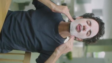 Erkek güzellik blogcusunun yüz maskelerini çıkarırken ve evde cilt bakımını çekerken kamera önünde konuşurken dikey görüntüsü.