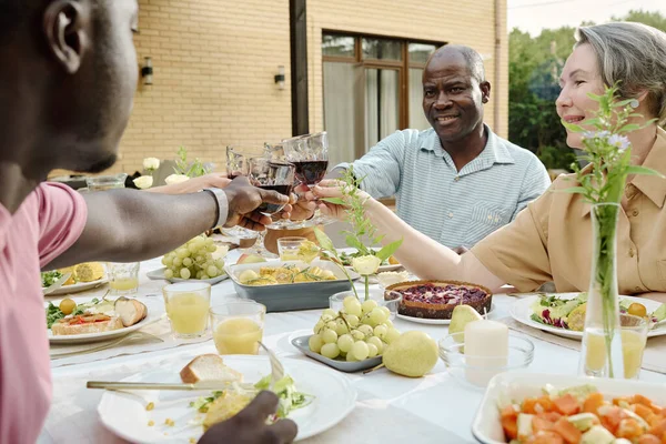 Happy mature couple with wineglasses toasting with their son and his wife while sitting by table served with homemade food during outdoor dinner