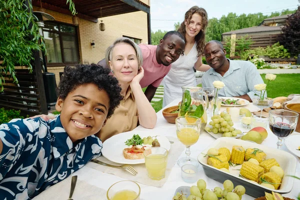 Cute African American boy in Hawaiian shirt taking selfie with his parents and grandparents while sitting by served table during family dinner