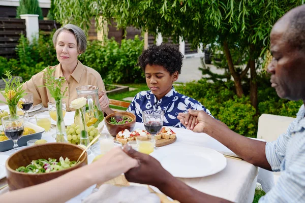 Cute little boy holding by hands of his grandparents during pray by table served with homemade food and drinks by outdoor family dinner