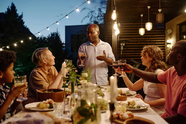Mature African American man with glass of red wine making toast while standing by served table in front of members of intercultural family