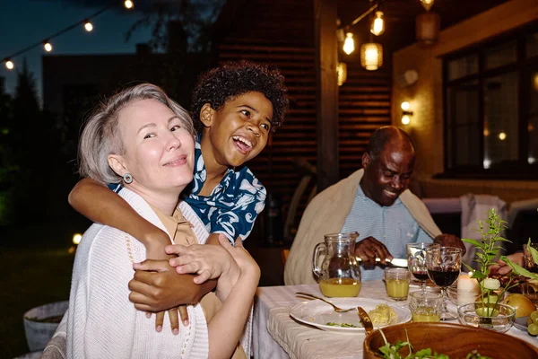 Happy mature woman looking at camera while her cute grandson embracing her by served table during family dinner at backyard