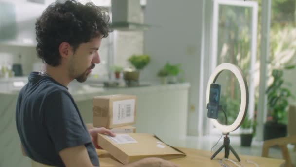 若い男は小包を解凍し サークルランプとスマートフォンの前でオンラインストアからの注文について話しながら 自宅でレビューVogを撮影 — ストック動画