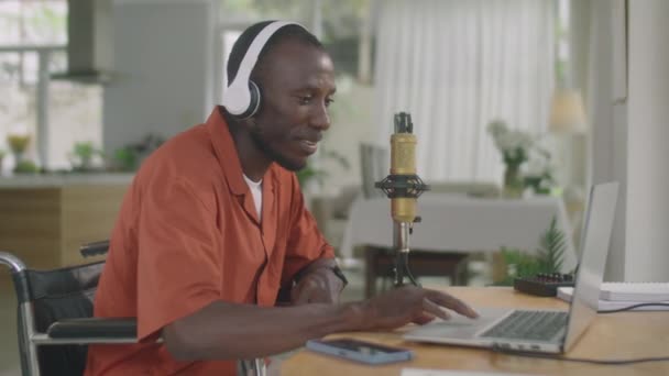 車椅子に座って自宅でポッドキャストを録音しながら マイクで話すワイヤレスヘッドフォンの障害を持つ若いアフリカ系アメリカ人の中型ショット — ストック動画