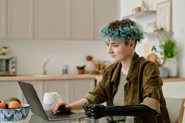 Young woman with prosthetic arm working on laptop while sitting in the kitchen