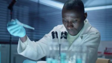 Steril eldivenli ve laboratuvar önlüklü Afrikalı Amerikalı bilim adamı laboratuvarda çalışırken mikroskop altında kimyasalları inceliyor.