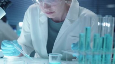 Steril eldivenli, koruyucu gözlüklü ve beyaz önlüklü kadın kimyager laboratuvarda deney yaparken mikroskop altında kimyasalları inceliyor.