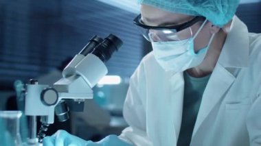 Tek kullanımlık eldivenli, şapkalı, maskeli ve koruyucu gözlüklü kadın bilim adamı mikroskoptan bakıyor ve laboratuvarda çalışırken notlar alıyor.