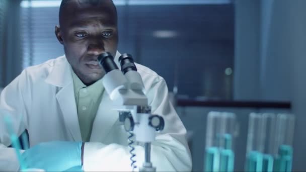 使い捨て手袋とラボコートのアフリカ系アメリカ人の科学者は 夜に実験室で働いている間に顕微鏡を通して見ています — ストック動画