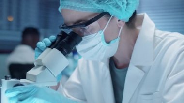Steril eldivenli, şapkalı, maskeli ve güvenlik gözlüklü kadın bilim adamının laboratuarda çalışırken mikroskoptan bakarken yakın plan çekimleri.