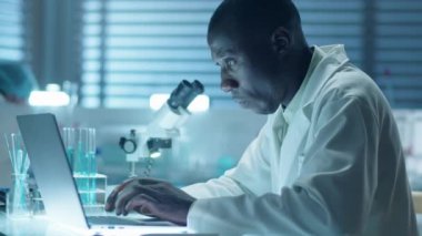 Laboratuvarda çalışırken, laboratuvar önlüklü Afrikalı Amerikalı erkek bilim adamının laptopla orta ölçekli görüntüsü.