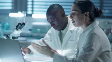 Genç beyaz kadın bilim adamının laboratuarda birlikte çalışırken Afrikalı Amerikalı meslektaşıyla dizüstü bilgisayarda bir şey tartışırken orta boy fotoğrafı.