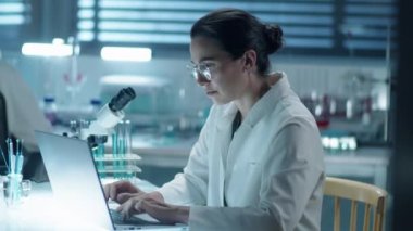 Laboratuvarda çalışırken laboratuvar önlüklü bir kadın kimyagerin laptopta yazı yazarken orta boy fotoğrafı.