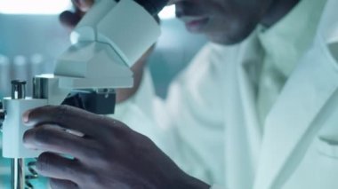 Laboratuvarda araştırma yaparken laboratuvar önlüklü Afrikalı Amerikalı erkek bilim adamının bileşik mikroskop topuzunu döndürdüğü seçici bir fotoğraf.