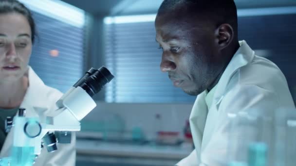白种人女科学家在一起做实验室研究时 一边做笔记 一边通过显微镜与非裔美国男性同事交谈 — 图库视频影像