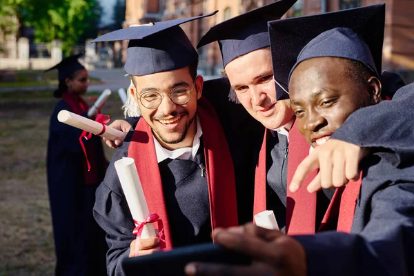 穿着毕业礼服的快乐男人们在智能手机上为自己画像 而不是在大学的户外 — 图库照片