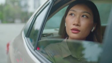 Kablosuz kulaklıklı Asyalı bir kadının arabasının arka koltuğunda otururken ve taksiye binerken açık pencereden bakarken görüntüsü.
