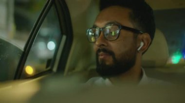 Kablosuz kulaklıklı sakallı bir iş adamının gece taksideyken açık araba camından bakarken çekilmiş göğsü.