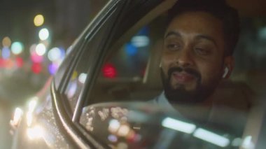 Kablosuz kulaklıklı bir iş adamının arabasının dışından, açık pencereden bakarken ve taksi sürerken gece şehrinin manzarasının keyfini çıkarırken gülümserken.