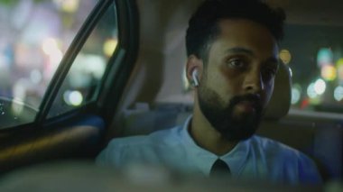 Kablosuz kulaklıklı ve resmi kıyafetli bir işadamı arabanın arka koltuğunda oturuyor ve gece taksi sürerken pencereden bakıyor.