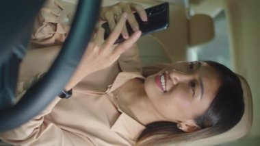 Genç Asyalı iş kadınının arabada oturduğu ve akıllı telefondan mesaj atarken gülümsediği dikey bir fotoğraf.