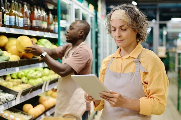 Saleswoman in uniform controlling goods online on tablet pc during her work in supermarket