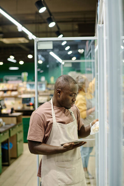 Афроамериканский работник магазина проверяет срок годности товаров с помощью планшетного ПК, находясь в супермаркете