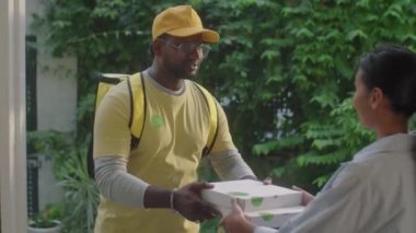 Afro-Amerikan erkek kurye kadın müşteriye pizza kutuları veriyor ve evde yemek dağıtırken onunla konuşuyor.