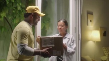 Asyalı kadın kapıyı açıyor, paket kutusunu kuryeden alıyor ve evde online sipariş alırken teslimat belgelerini imzalıyor.