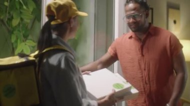 Neşeli Afro-Amerikalı adam yemek servisi kullanırken kadın kuryeden pizza kutuları alıyor.