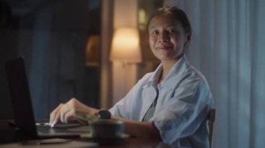 Bilgisayarı kullanan Asyalı bir kadının portresi ve akşam evde çalışırken kameraya gülümseyerek poz vermesi.