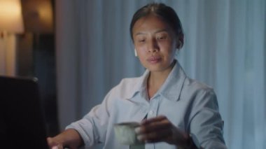 Kablosuz kulaklıklı Asyalı kadın dizüstü bilgisayar kullanıyor, not alıyor ve geceleri karanlık oturma odasında çalışırken çay içiyor.