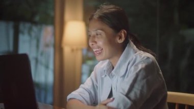 Neşeli Asyalı kadın, akşam vakti evdeki karanlık oturma odasında dizüstü bilgisayarla gülümseyip sohbet ediyor.