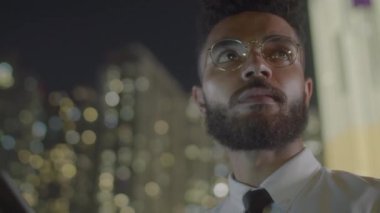 Düşük açılı, resmi kıyafet ve gözlüklü genç iş adamının gece vakti şehrin dışında dikilirken akıllı telefon kullanarak etrafı kolaçan ederken görüntüsü.