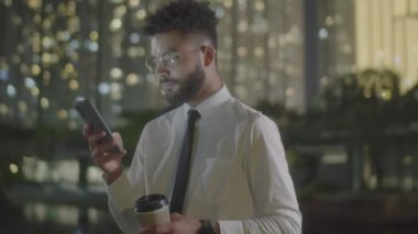 Genç sakallı iş adamının gece vakti şehirde dikilmiş elinde kahve fincanı tutarken ve akıllı telefondan mesaj yazarken orta boy fotoğrafı.