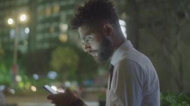 Formalite icabı genç işadamının gece sokakta oturduğu ve akıllı telefondaki mobil interneti taradığı orta ölçekli bir yan görüntü.