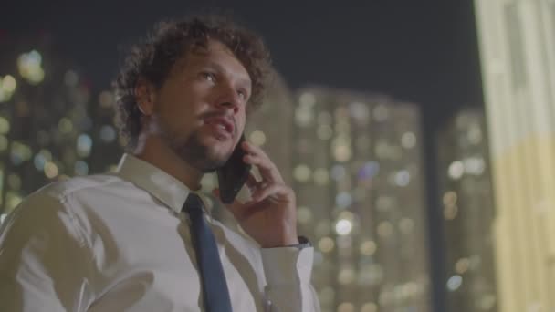 屋外に立って ナイトシティで携帯電話で話す正式な衣装でコーカサス人のビジネスマンの低角度ショットをチェスト — ストック動画