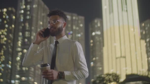 夜に街の屋外で立っているフォーマルウェアの若いビジネスマンの中程度の低角度ショット 使い捨てコーヒーカップを保持し 携帯電話で話す — ストック動画