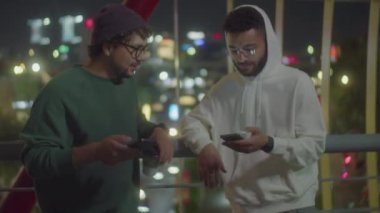 Şehirdeki yaya köprüsünde dikilen ve büyük şehirde gece vakti akıllı telefonlarıyla bir şey tartışan sokak tarzı kıyafetler giymiş iki erkek arkadaşın orta boy fotoğrafı.