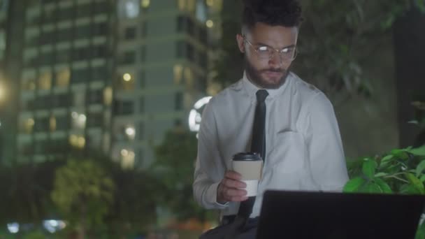 夜の街のベンチに座って コーヒーカップに行ってラップトップでオンラインで作業するビジネスマンのミディアムショット — ストック動画