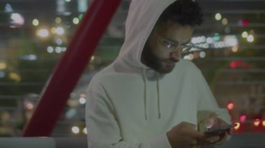 Kapüşonlu genç adamın gece yarısı yaya köprüsünde demiryolu kenarında durup akıllı telefondan mesaj yazarken orta boy fotoğrafı.