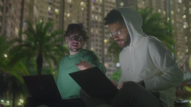 説明公園に座っているカジュアルウェアの2人の若い男性フリーランサー ラップトップでプロジェクトを議論し 都市で夜間にオンラインで作業しながら公共のインターネットを使用する — ストック動画