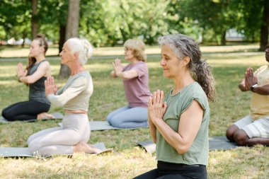 Kıdemli kadın gözleri kapalı bir şekilde parkta oturmuş, diğer insanlarla birlikte meditasyon yapıyor.