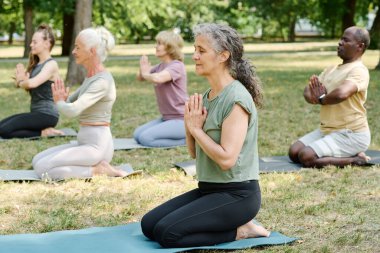 Sağlıklı bir kadın egzersiz minderinde oturur ve gözleri kapalı meditasyon yapar. Arka planda başkaları da vardır.