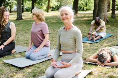 Egzersiz minderinde oturan ve kameraya gülümseyen yaşlı kadının portresi, parkta yoga yapıyor.