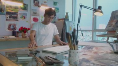 Sanat stüdyosunda yürüyen genç Asyalı adam, masada oturuyor ve albümde çizim yaparken kurşun kalem kullanıyor.