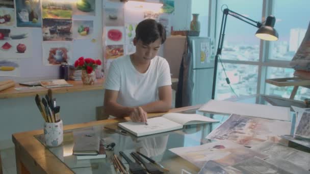 クリエイティブスタジオでデスクで働いている間にアルバムにワックスクレヨンで絵を描く若いアジア人アーティストのショットでズーム — ストック動画