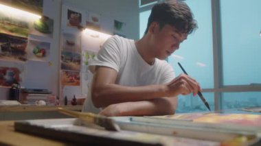 Resim stüdyosundaki masada resim çizerken genç Asyalı ressamın boya fırçası kullandığı orta boy bir fotoğraf.
